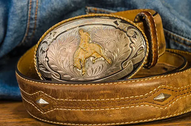 Vintage silver buckle with cowboy on bucking bronc.  Leather belt with studs against blue denim work shirt background.