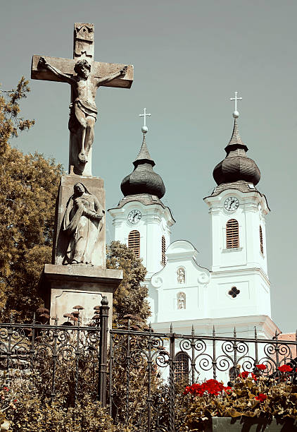 Budapest, Hungary, April 2025The facade of the Pest Franciscan Church.The upper facade of a Baroque-style church features detailed white and pale yellow stucco architecture with sculpted statues of religious figures, including a saint with a crosier and another with a sword, positioned on pedestals. A prominent cross rises from the roofline, flanked by ornate urns and a radiant sunburst motif. Below, a niche contains a relief sculpture of the crucifixion scene with Jesus on the cross. The structure includes arched windows and decorative scrolls, set against a cloudy gray sky during daytime. No people are present. This image captures the grandeur of historic religious architecture in Europe, suitable for commercial use in travel magazines, architectural books, historical documentaries, religious publications, and tourism promotions highlighting cultural heritage sites.