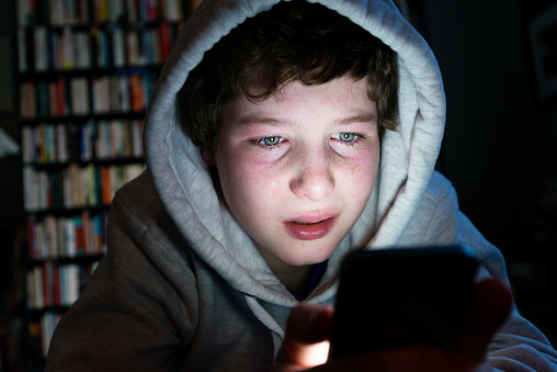 Candid colour portrait of a young teen in tears staring intently into a smart phone. His face is illuminated by the light from the digital device that he is holding, he is wearing a grey hoodie with the hood up and he has a down beat look on his face, maybe he is being bullied online or looking at something more sinister online. Horizontal format with a dark background that could be suitable for some copy space.