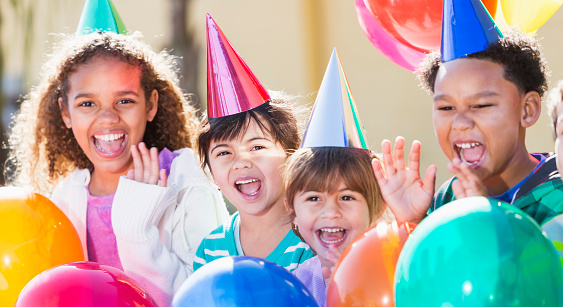 A group of four multi-ethnic children having fun at a birthday party laughing and waving, surrounded by colorful balloons and wearing party hats.  They are mixed ages from 4 to 9 years old.  The main focus is on the little girl wearing a pink hat.