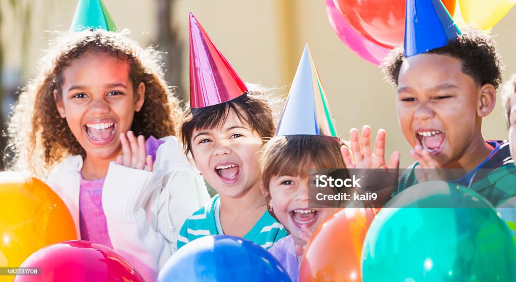 Multiracial children at a birthday party A group of four multi-ethnic children having fun at a birthday party laughing and waving, surrounded by colorful balloons and wearing party hats.  They are mixed ages from 4 to 9 years old.  The main focus is on the little girl wearing a pink hat. Child Stock Photo