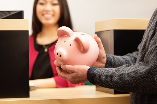 A senior man with retirement fund in piggy bank in front of bank teller at the bank counter window. The hands is holding the piggy bank with the banker bank teller in the background. Photographed in horizontal format.