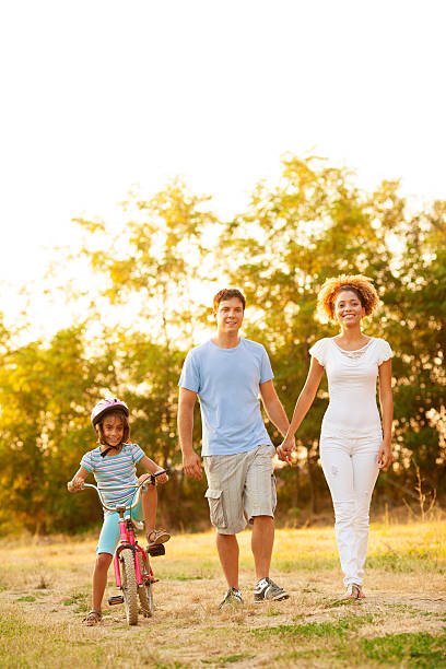 Mixed race family enjoy walking outdoors, little girl riding a bicycle. Looking at camera and smiling.