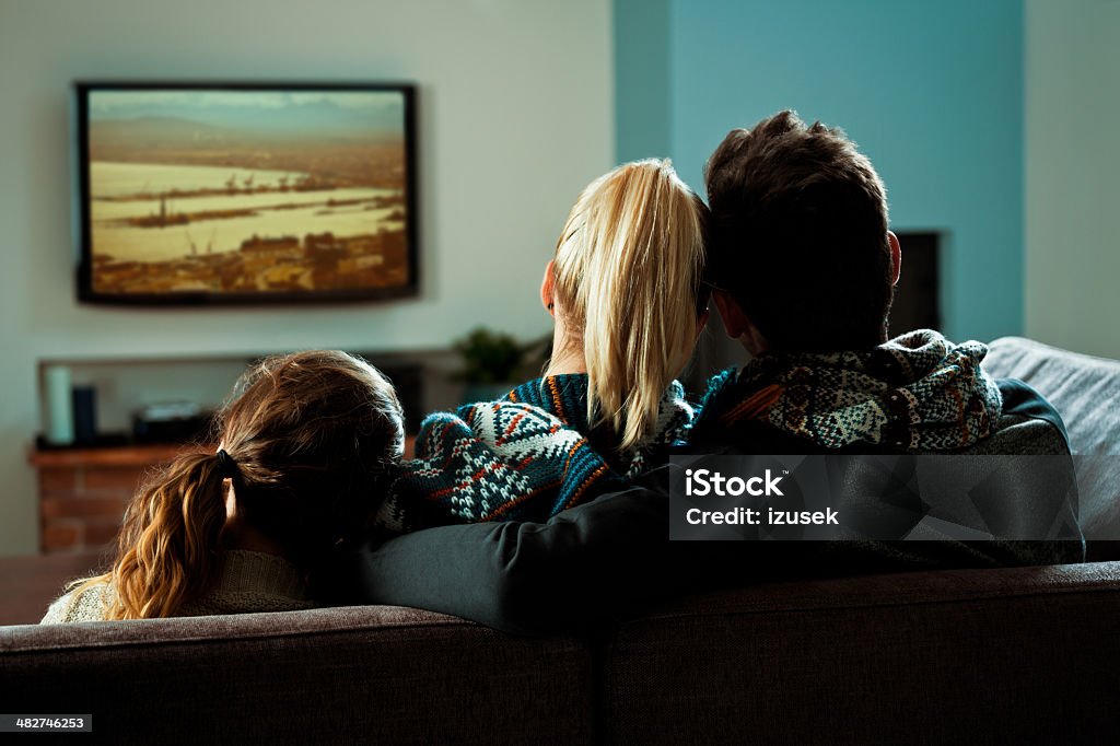Family watching TV Back view of family sitting on sofa at home and watching TV. Dark tones. Television Set Stock Photo