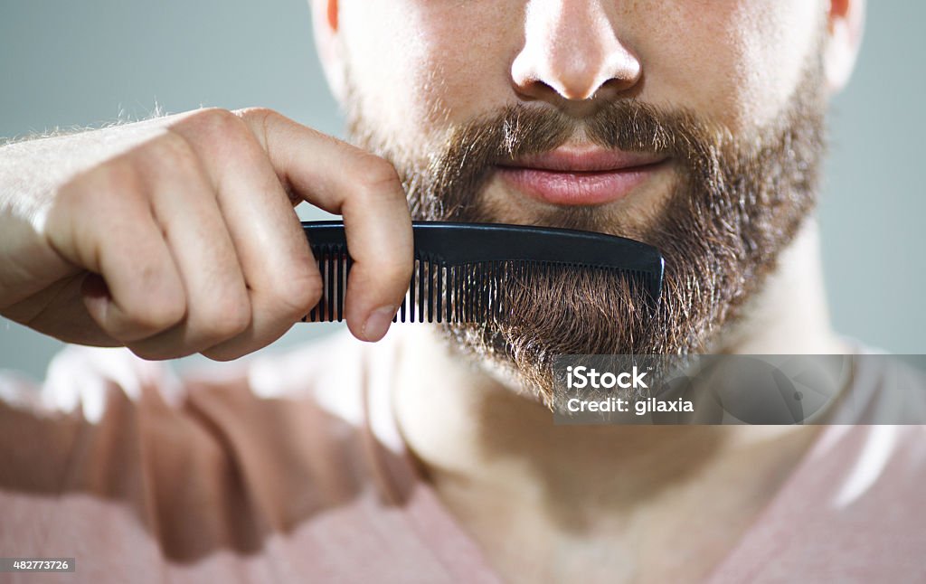 Unrecognizable man combing his beard Closeup of unrecognizable caucasian man combing his beard and mustach with small black comb.He has neat,fully grown brown beard and mustache.Studio shot over gray background. ,=Front view. Beard Stock Photo