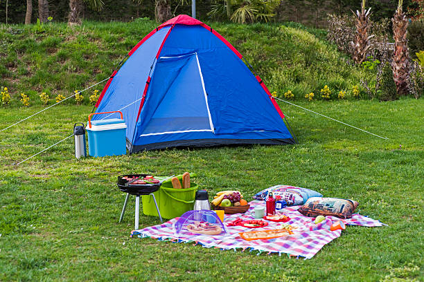 Two teenage girls sit on their mat and talk cheerfully, one holding a drink bottle as they enjoy the peaceful surroundings and gentle light of their campsite.