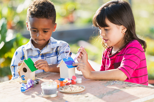 Two multi-ethnic children sitting outdoors at a table in a garden on a sunny day, doing an arts and crafts project, painting little bird houses.  The African American boy and mixed race Hispanic girl are both 5 years old.  They are serious expressions on their faces, concentrating on their projects.