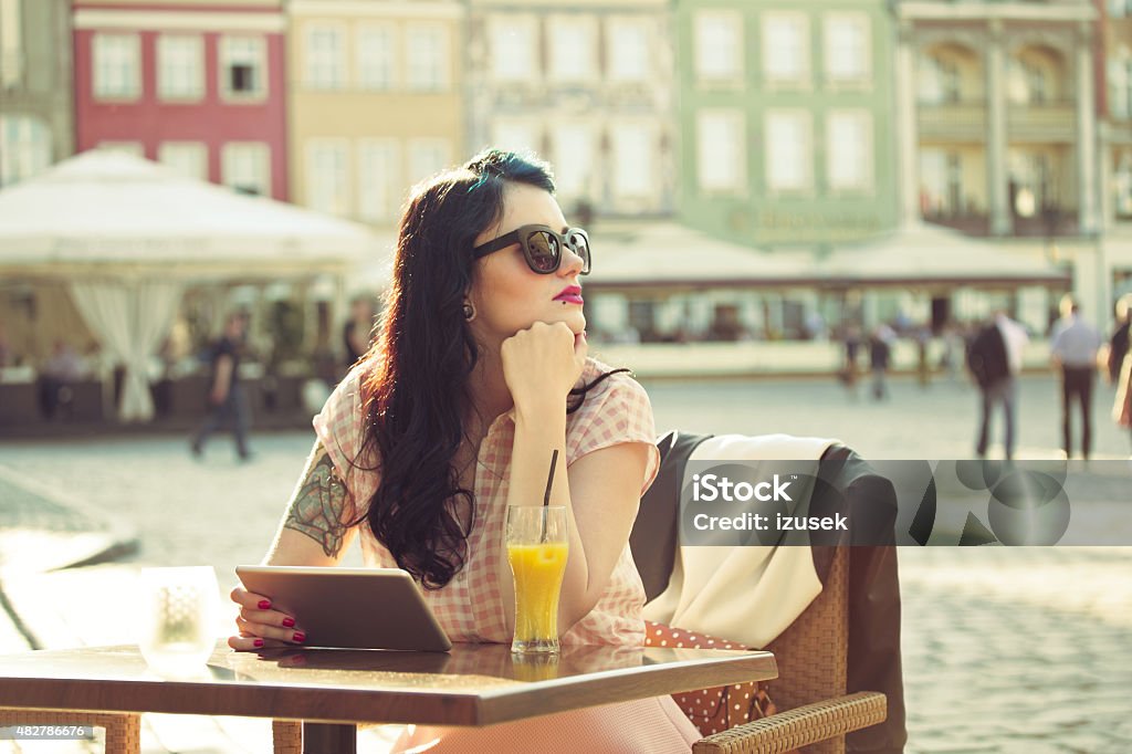 Young woman using a digital tablet in the outdoor restaurant Young hipster woman with tattoo on her arm wearing sunglasses sitting at the table in the outdoor restaurant in the city, holding a digital tablet and looking away. Orange juice on the table. Summer time.  Asking Stock Photo