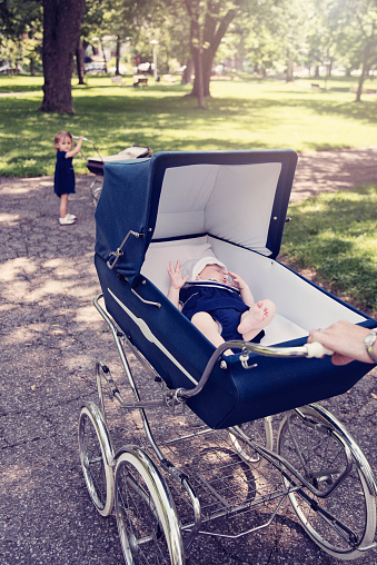 Mother and little daughter are proudly walking theirs babies in original vintage prams. Close-up on the baby sleeping. Dressed in classic dresses in white and navy blue, they are in a public city park, in summer. Baby is 5 month, little girl is two. Trees in the background. Vertical.