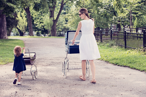 Mother and little daughter are proudly walking theirs babies in original vintage prams. Dressed in classic dresses in white and navy blue, they are in a public city park, in summer. Baby is 5 month, little girl is two. Trees in the background. Full length horizontal shot with copy space.