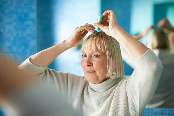 senior woman checking hairline for hair loss old caucasian woman applying lotion to prevent hair loss, looking at mirror in bathroom woman hairline stock pictures, royalty-free photos & images