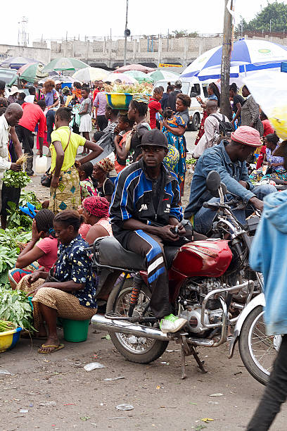 Motorcycle Taxi in DRC, AFRICA Kinshasa, Democratic Republic of the Congo - June 10, 2015: Two men seating on Chinese motorcycles at a market in UPN around them people are selling, buying and walking around. There are women all around selling green leaves and vegetables. In the background, there's some construction work. kinshasa stock pictures, royalty-free photos & images