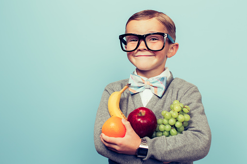 A young nerd boy with glasses is excited to eat his fruit. He is holding a bunch of fruit and has an excited expression on his face. 