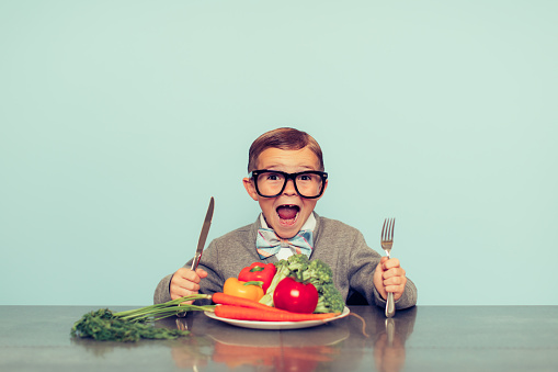 A young nerd boy with glasses is excited to eat his vegetables. He is sitting at a table and has an excited expression on his face. 