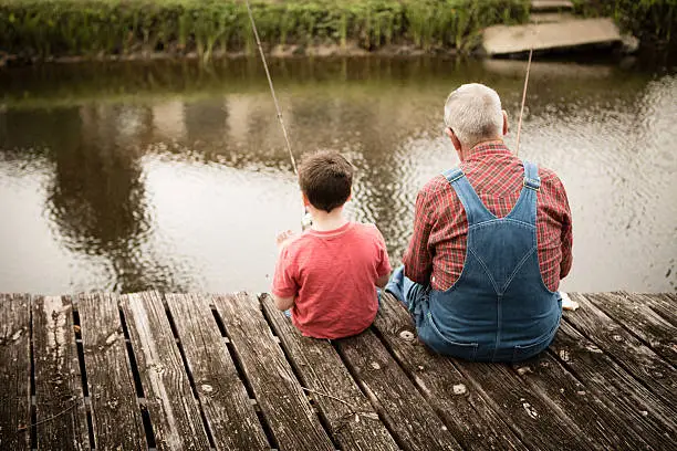 Color image of a senior man sitting on an old, wooden dock while fishing with his young great grandson.
