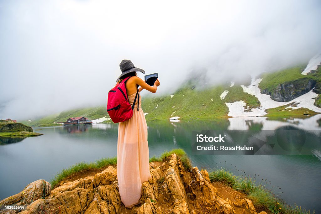 Traveler on the mountain lake Female traveler in long dress and big hat with red backpack photographing mountain lake with fog and snow. Traveling in Romania 2015 Stock Photo