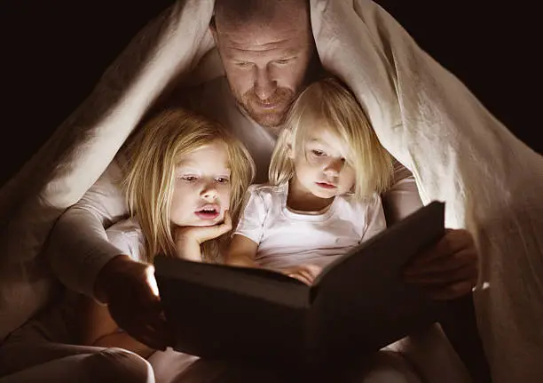Shaved middle aged father and young blonde daughters reading a book together at bedtime