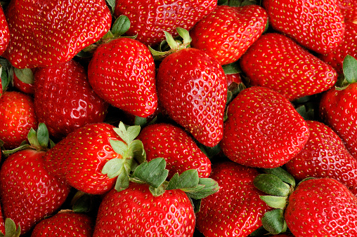 A full-framed background filled with fresh, ripe strawberries.  The strawberries are a bright red color with green stems and leaves.  The fruit are in a good condition with details of small yellow seeds on each strawberry.  The strawberries are piled on top of one another.