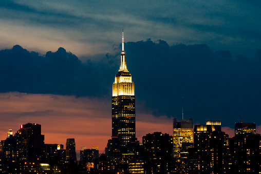 This is a horizontal, color, royalty free stock photograph shot with a Nikon D800 DSLR camera. It is an early summer evening in New York City. The clouds above the dense urban metropolis are turning colors as sunset dusk is approaching. The Empire state Building is visible in the background. The city is becoming illuminated from the glowing windows. Photographed from a high angle view across the East River from Williamsburg, Brooklyn.