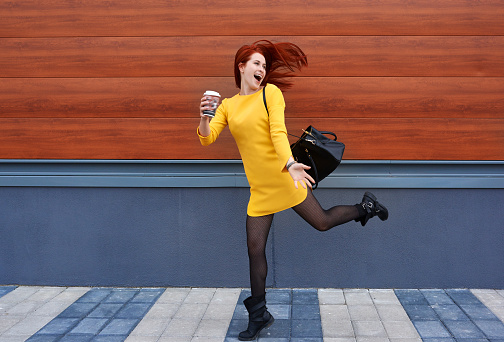 front view of woman in yellow dress holding coffee and jumping, dancing and starting her day with energy.