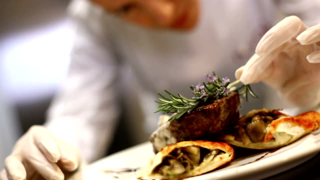 Professional female chef placing finishing touches by putting rosemary on steak just before serving. She's wearing protective gloves when touching ready to eat food.HD 1080