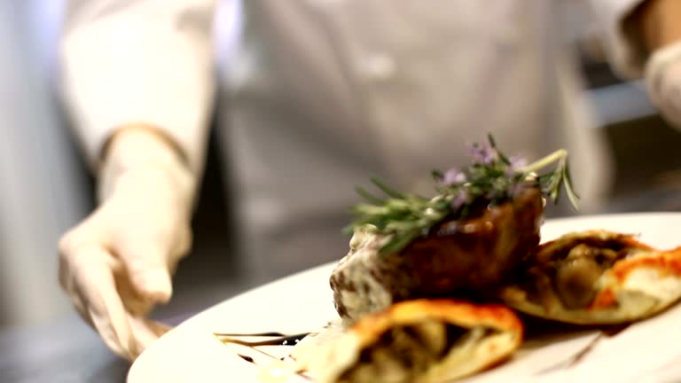 Professional female chef placing finishing touches by putting rosemary on steak just before serving. She's wearing protective gloves when touching ready to eat food.HD 1080