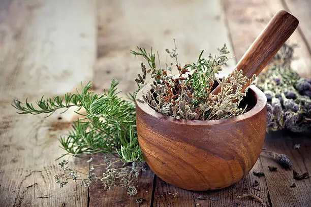Fresh herbs inside a wooden mortar and pestle bowl on a wooden plank table with knotholes and nails.  The herbs have multiple colors and textures.  The bowl has a swirl pattern and is surrounded by piles of additional fresh herbs.