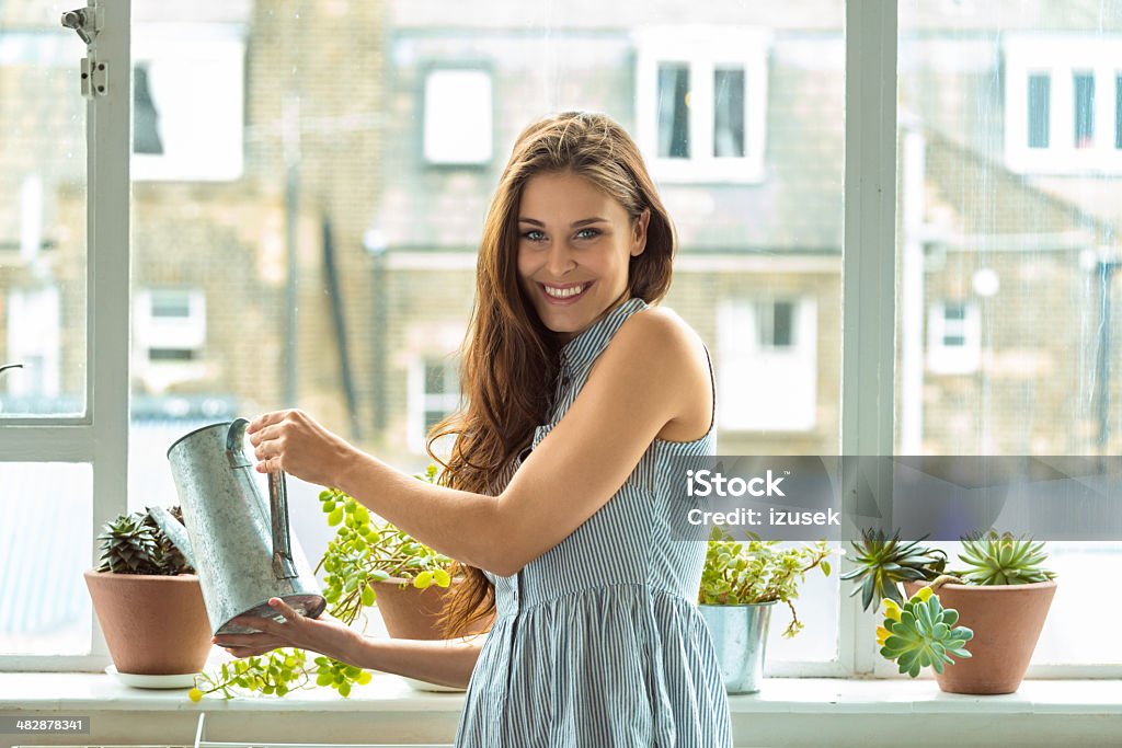Woman watering plants Beautiful young woman watering potted plants at her home, smiling at camera. One Woman Only Stock Photo