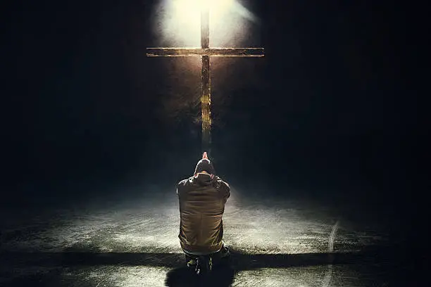 A man worships in prayer, his hands folded as he kneels in front of a crucifix in a dark grunge setting.  A single light illuminates the scene from above and behind.  Imagery intended to represent the crucifixion and resurrection of Jesus Christ celebrated on Easter sunday.  Horizontal image with copy space.