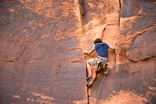 Athletic young man artfully scaling a sheer red rock sandstone wall near Moab Utah.