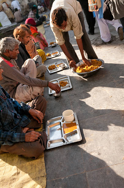 Udaipur, India - January 7, 2011: Poor people are receiving a free meal at the local Shree Jagdish Hindu temple in Udaipur, India. A man is distributing lentils and rice cooked together on metal plates. The homeless people are waiting for food sitting in a row. In India, poor people such as these elderly people do not have access to social security. They have to rely on their local community. Local temples collect donations to feed the starving people who are living in the streets. Some temples such as this also provides free medical service to those in need.