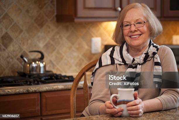 Smiling Senior Woman Sitting With Cup In Hand In Kitchen Stock Photo - Download Image Now