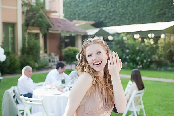 Photo of Woman standing at outdoor party with flowers showing off new ring smiling