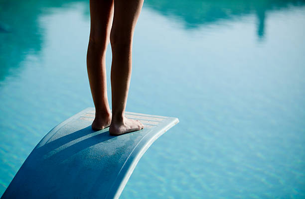 Shot of bare legs on diving board above blue water A person standing on a diving board.  Only the back of the person's legs and feet are visible.  The person is standing up straight.  There's a light reflecting off of the person's feet and the top of the board.  The diving board bends towards the water underneath the person's weight.  The diving board is light colored and blends in a little with the water.  The board is also textured with a raised border.  The water is light in color with small ripples.  Reflections of other objects in the water create a border at the top of the image. diving board stock pictures, royalty-free photos & images