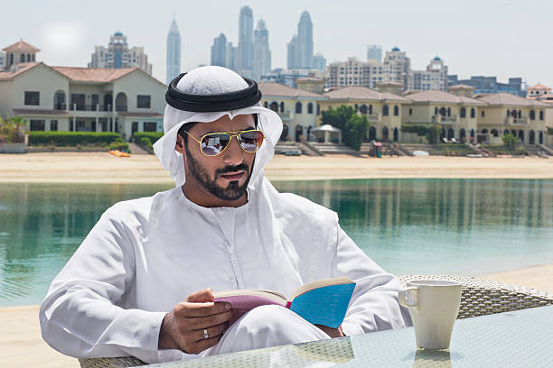 Relaxing in the terrace at home Middle Eastern man relaxing in the back yard reading a book with a cup of coffee, he's wearing a dish dasha. Dubai Marina is in the background. arabian sunglasses stock pictures, royalty-free photos & images