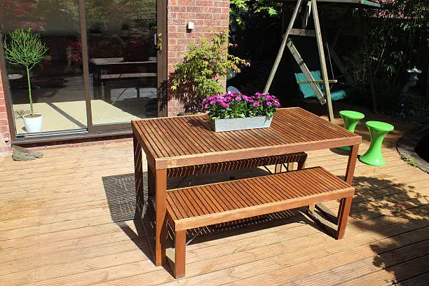 Photo showing a contemporary style, slatted wooden table and matching benches.  This stylish garden furniture is pictured standing on timber decking, outside the patio doors leading into the sunny living space at the back of the house.