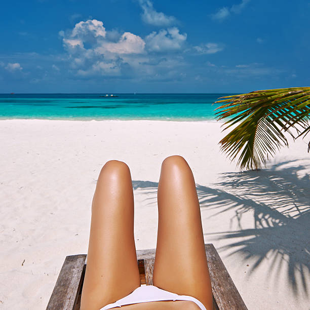 Summer holidays. Beautiful young woman in colorful swimsuit and white sarong and sunglasses listening to music in headphones and lying on back on white sand.