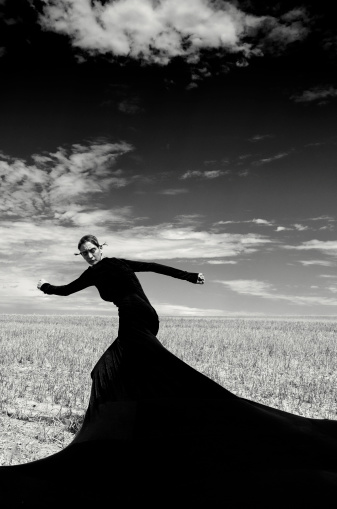 A surreal outlooking female human being posing with her long dress. She is trying to get away from something. Monochrome processed image with strong contrast and structure. Some grain visible.