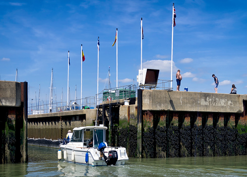 Shotley Gate, Suffolk, England - May 27, 2015: A small cabin cruiser, with people on board, entering the lock which connects the River Orwell estuary with Shotley Marina, in Suffolk, eastern England. The estuary is tidal so the lock is necessary to regulate the respective heights of the marina and the harbour. A few people are on the quayside, indulging in some line fishing.