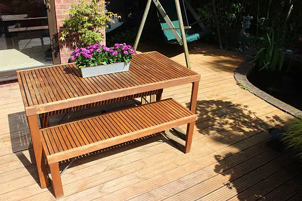 Photo showing some slatted wooden garden furniture, which is part of a sunny back garden.  The table and its two benches are positioned by the patio doors that lead into the house, providing a useful outdoor eating space.  A zinc trough of osteospermum flowers is sitting on the table.