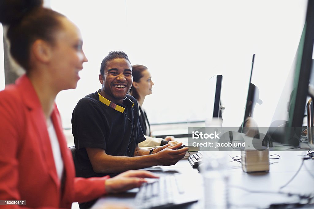 Young student in class with other students working on computers Young afro american man looking at camera smiling while working on computer in modern classroom. Young students sitting at college computer lab. Computer Lab Stock Photo