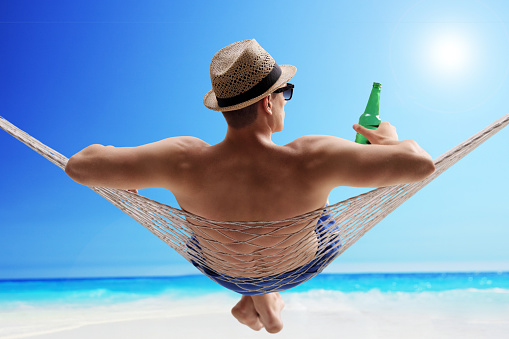 Relaxed young guy lying in a hammock and drinking beer on a sunny beach by the ocean