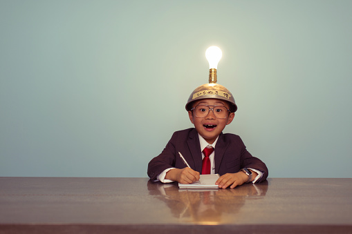 A young Japanese businessman sits at office desk searching for successful business ideas by using a thinking cap. He is working on ideas with the light bulb lit up. The image is retro styled with a blue background. They are wearing business suits and ties.