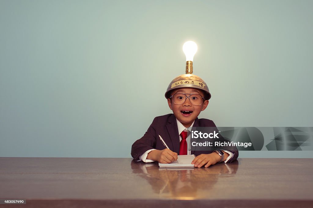 Surprised Japanese Business Boy Wearing Lit Up Thinking Cap A young Japanese businessman sits at office desk searching for successful business ideas by using a thinking cap. He is working on ideas with the light bulb lit up. The image is retro styled with a blue background. They are wearing business suits and ties. Inspiration Stock Photo
