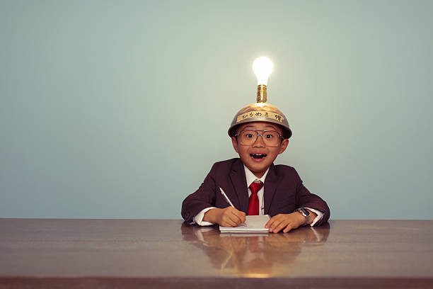 Surprised Japanese Business Boy Wearing Lit Up Thinking Cap A young Japanese businessman sits at office desk searching for successful business ideas by using a thinking cap. He is working on ideas with the light bulb lit up. The image is retro styled with a blue background. They are wearing business suits and ties. resourceful stock pictures, royalty-free photos & images