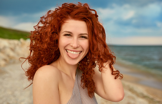 woman portrait on the beach, smiling and sticking out tongue, enjoying summer vacation.red curly hair.