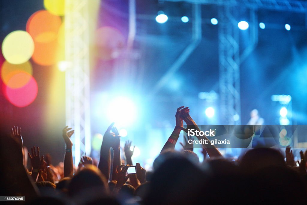Rock concert. Rear view of people cheering at concert during music festival.Large group of unrecognizable adults with their arms raised.Some of them holding beer cans.There's a stage and a band performing in background,out of focus.Stage is lit blue. Arms Outstretched Stock Photo
