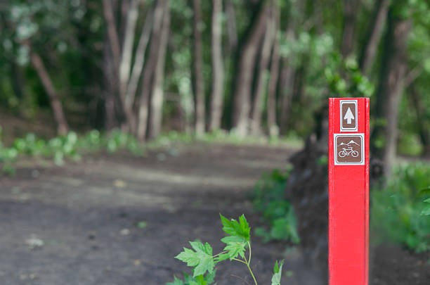 Red directional sign for bikes, leading into wooded area by park during summer.