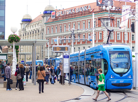 Zagreb, Сroacia - September 22, 2014: Streetcar station in the Zagreb central square Ban Jelacic. A blue Tram is just living the station; woman is crossing the treks while a lot of people wait at station for next tram. In background are houses with lot of commercial panels. It is grey autumn day.