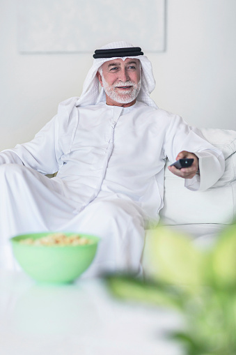 Emirati senior man watching television,he is changing television channel .